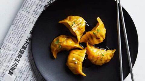 Five dumplings are arranged on a black plate with chopsticks, next to a bowl of chili sauce, a green bottle, and a newspaper with Chinese text—perfect for celebrating Lunar Chinese New Year.