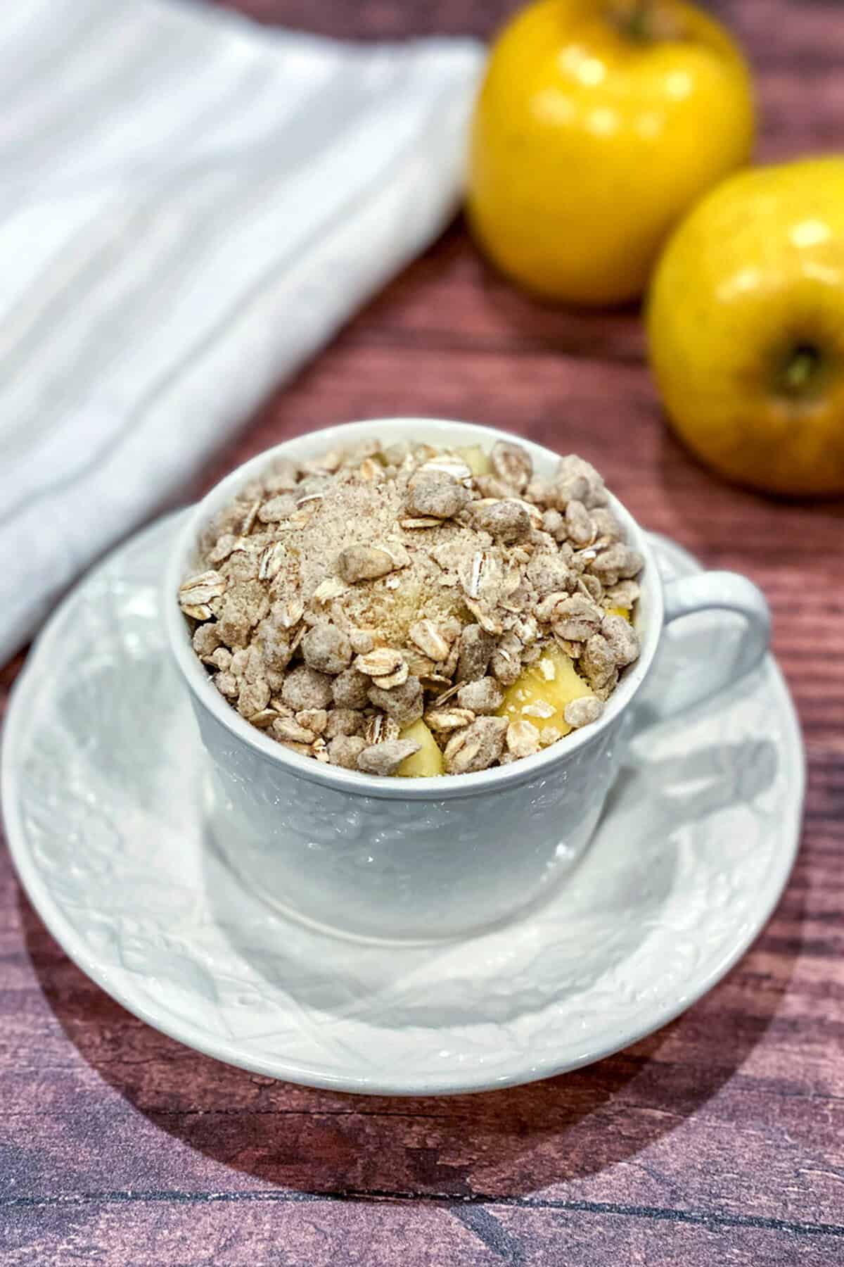 A white teacup filled with Apple Crisp in a Mug sits on a matching saucer atop a wooden surface. Two yellow apples and a striped cloth are in the background.