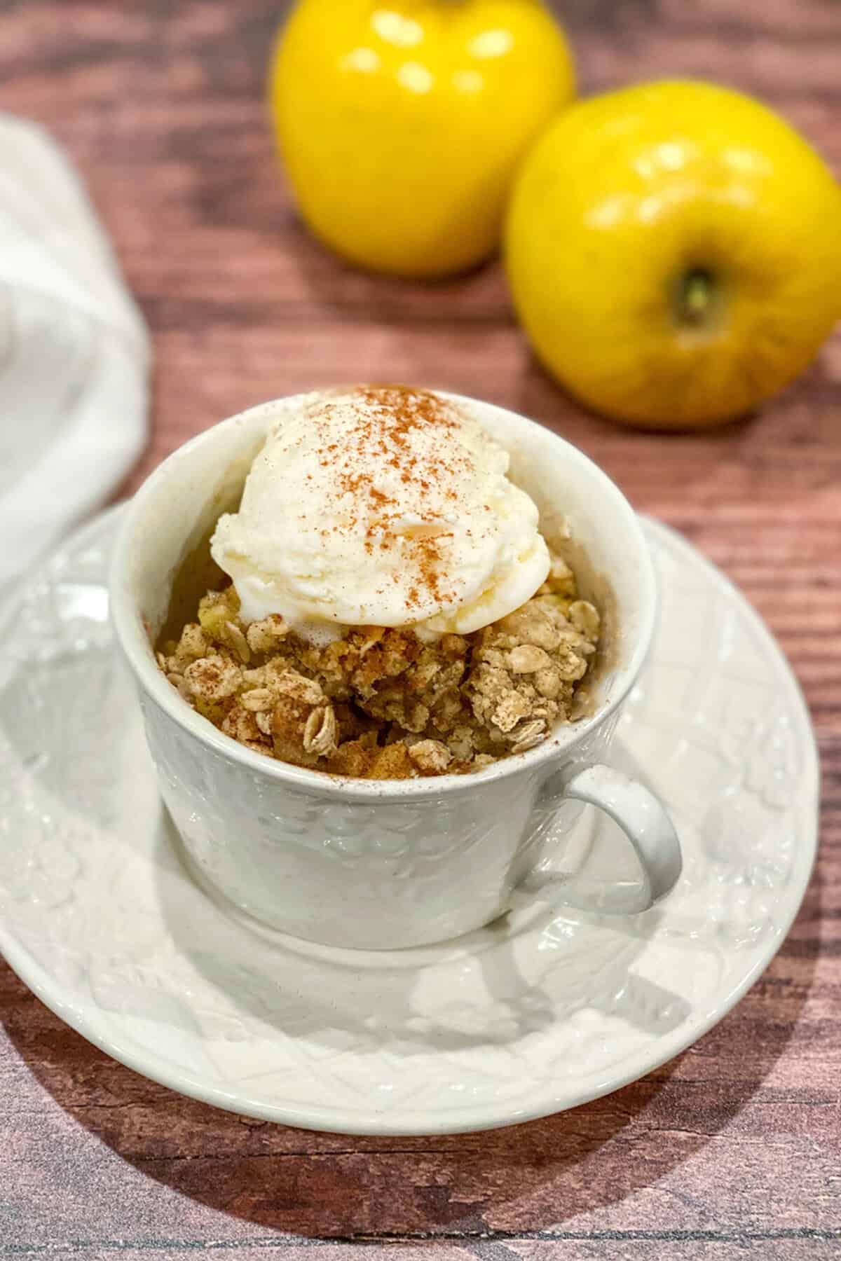 A white teacup serves up Apple Crisp in a Mug, topped with vanilla ice cream and a sprinkle of cinnamon, resting on a saucer with two yellow apples in the background on a wooden surface.