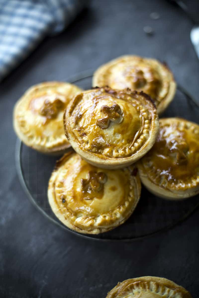 A stack of small, round, golden-brown meat pies-reminiscent of classic Irish pub recipes-sits on a wire rack, with a textured dark surface and a blue-checkered cloth in the background.