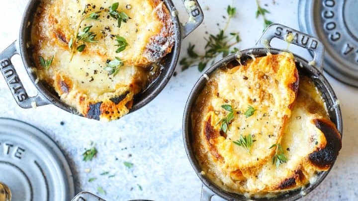Four mini cast iron pots filled with French onion soup, a classic among French recipes, topped with toasted bread and melted cheese, garnished with fresh herbs, placed on a light surface with bread slices nearby.