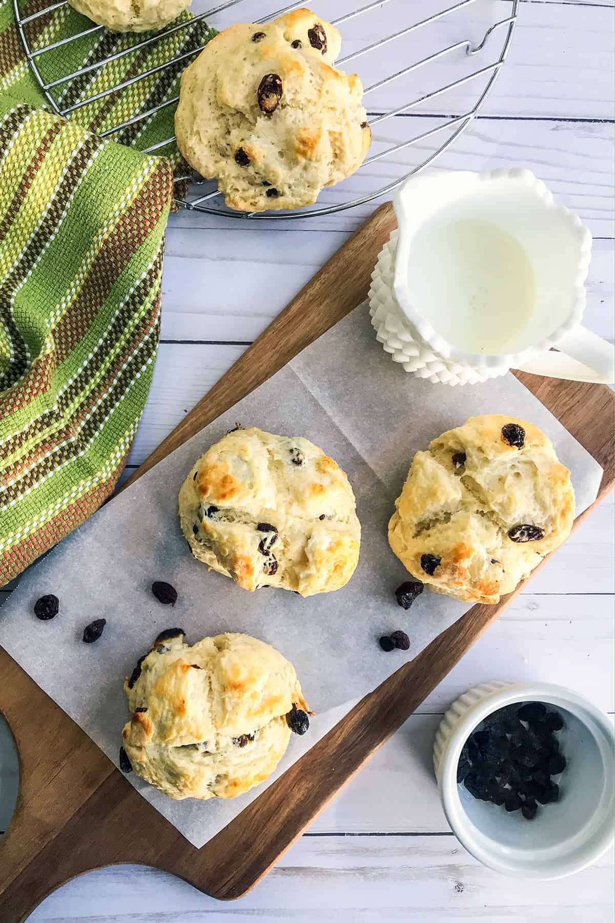 Mini Irish Soda Bread rest on a parchment-lined wooden board, with a small bowl of raisins, honey, and a glass jug of milk.