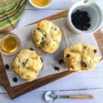 Three raisin scones on parchment paper atop a wooden board, with a bowl of raisins, a glass of honey, and a colorful towel nearby—perfect alongside mini Irish soda bread for a cozy teatime treat.