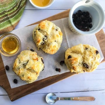 Three raisin scones on parchment paper atop a wooden board, with a bowl of raisins, a glass of honey, and a colorful towel nearby-perfect alongside mini Irish soda bread for a cozy teatime treat.