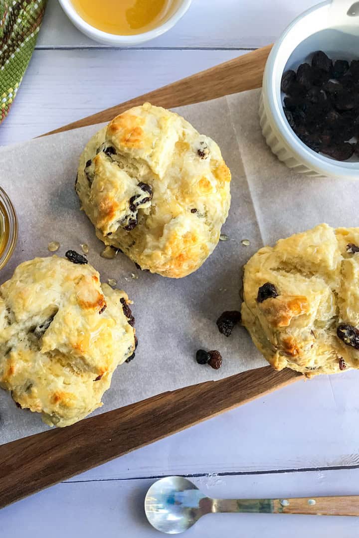 Three raisin scones on parchment paper atop a wooden board, with a bowl of raisins, a glass of honey, and a colorful towel nearby—perfect alongside mini Irish soda bread for a cozy teatime treat.