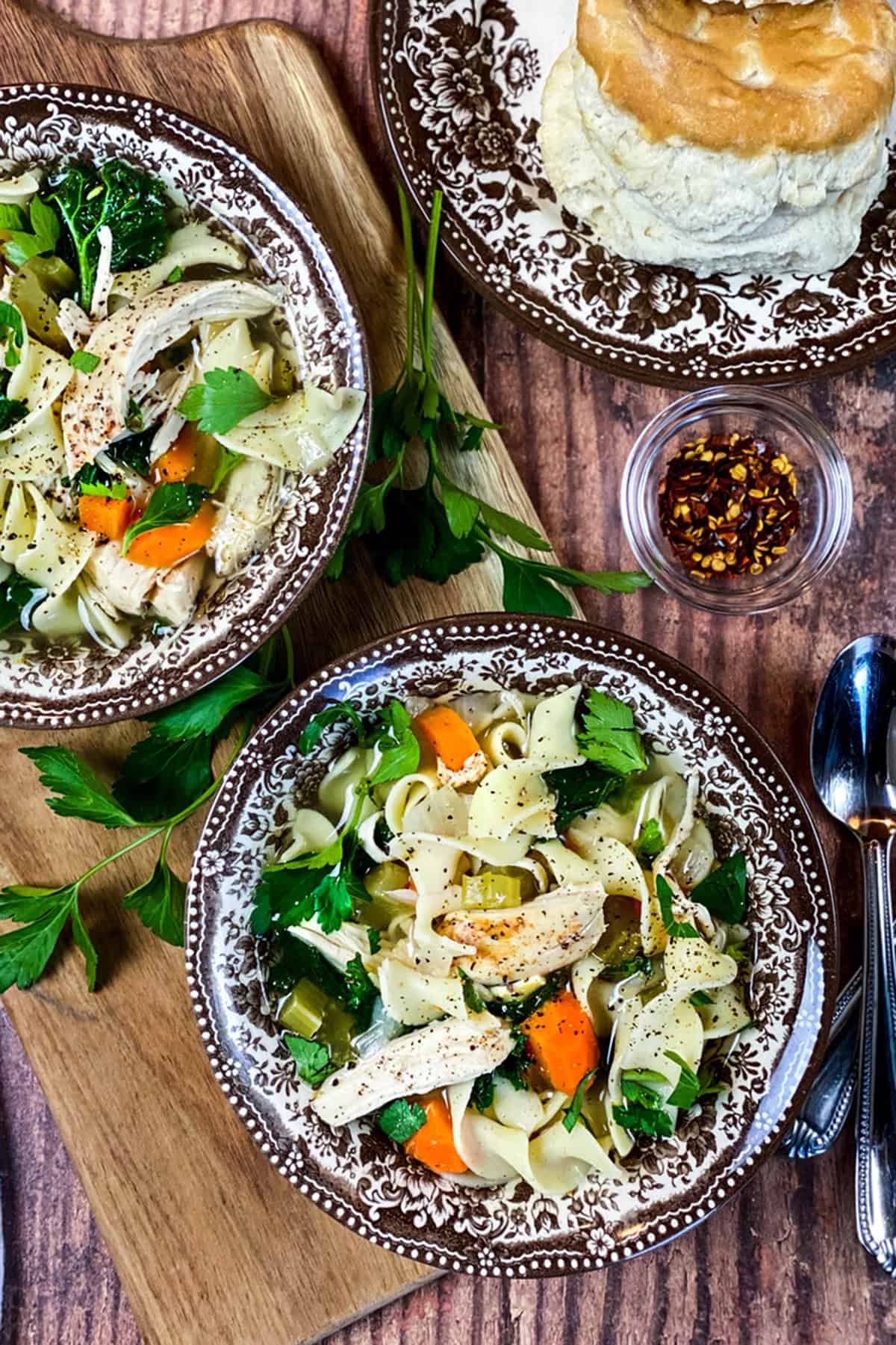Two bowls of slow cooker chicken noodle soup on a wooden serving board with fresh parsley on the side.