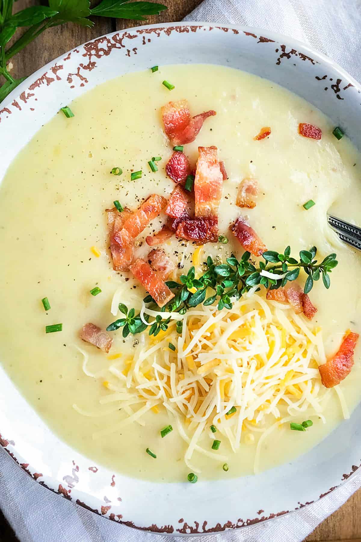 A bowl of creamy Slow Cooker Potato Soup topped with shredded cheese, bacon pieces, chopped chives, and fresh thyme.