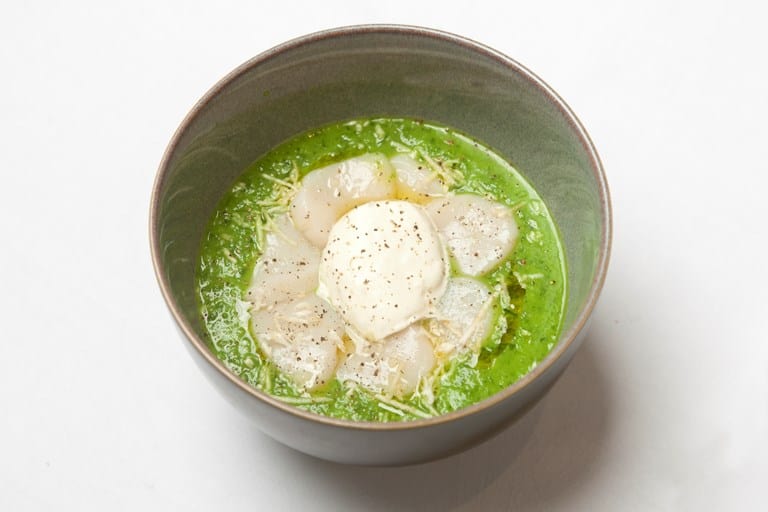 A bowl of green soup inspired by Irish pub recipes, topped with sliced scallops, shredded cheese, and a poached egg, garnished with black pepper.