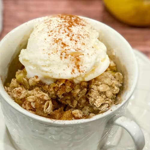 A white mug filled with Apple Crisp in a Mug, topped with a scoop of vanilla ice cream and a sprinkle of cinnamon, sits on a white saucer against a wooden background.