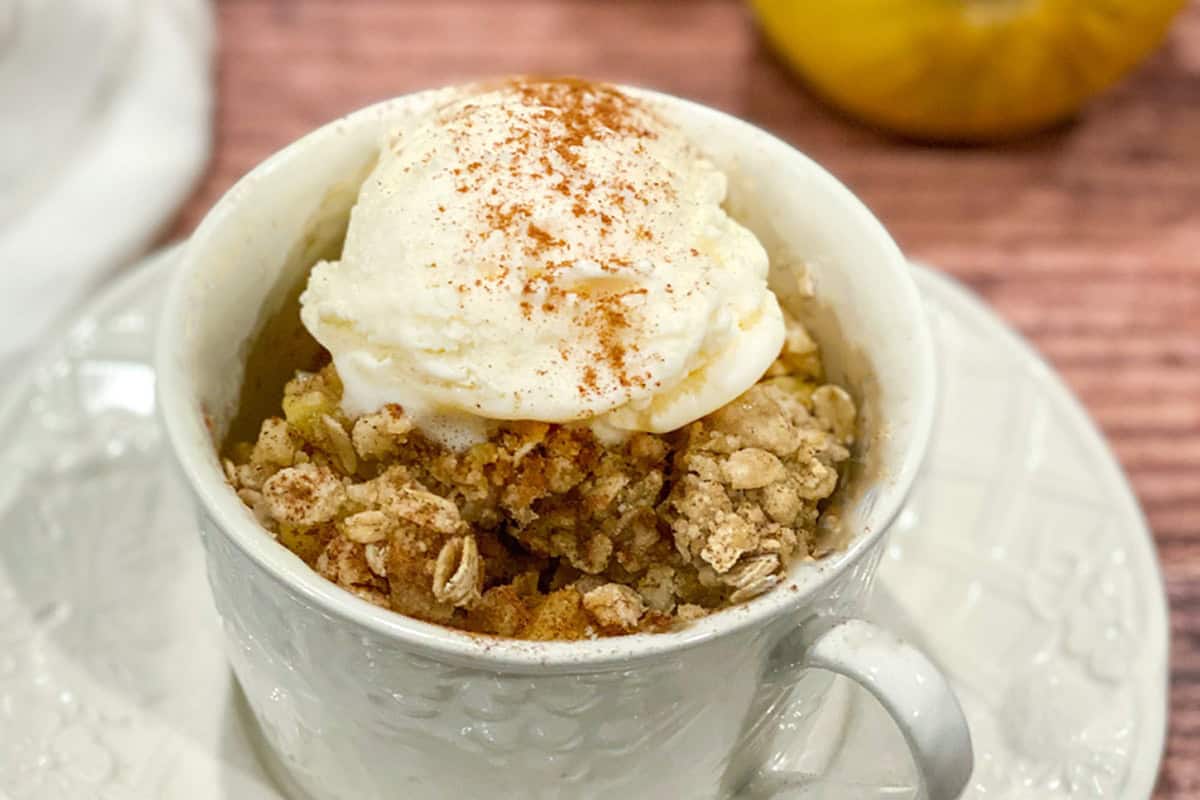 A white mug filled with Apple Crisp in a Mug, topped with a scoop of vanilla ice cream and a sprinkle of cinnamon, sits on a white saucer against a wooden background.