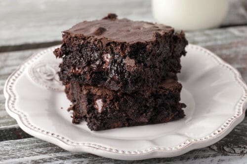 Two thick, fudgy brownies with melty chocolate chunks are stacked on a decorative white plate, placed on a rustic wooden surface, showcasing the ultimate indulgence for fudgy brownie lovers.