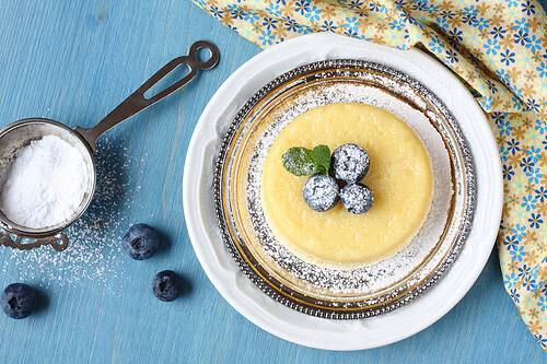 A small cheesecake, reminiscent of lemon pudding cakes, topped with three blueberries and a mint leaf, dusted with powdered sugar on a glass plate with a white rim, sits next to a sifter and blueberries on a blue surface.