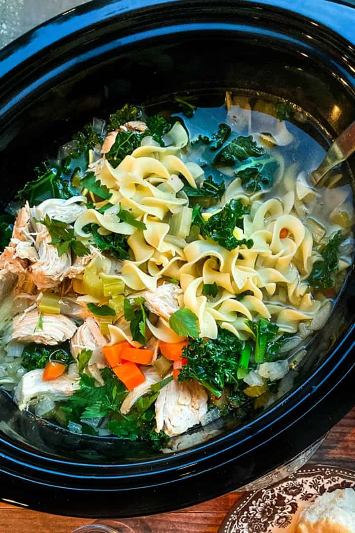 Chicken Noodle Soup with egg noodles and greens in a black slow cooker insert ready for serving.