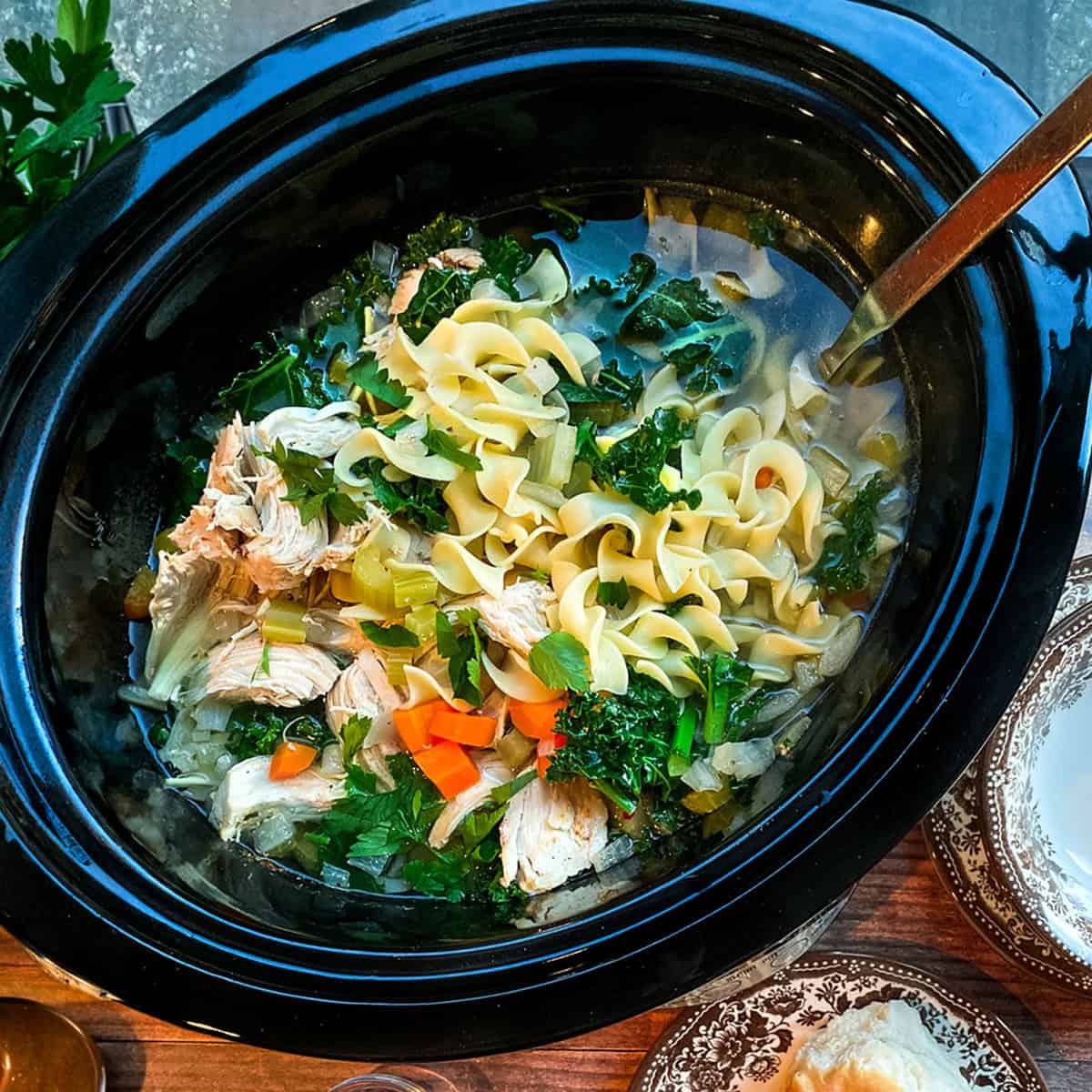 Chicken Noodle Soup with egg noodles and greens in a black slow cooker insert ready for serving.