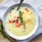 A bowl of creamy Slow Cooker Potato Soup topped with shredded cheese, crispy bacon, fresh herbs, and chives, with a spoon resting inside.
