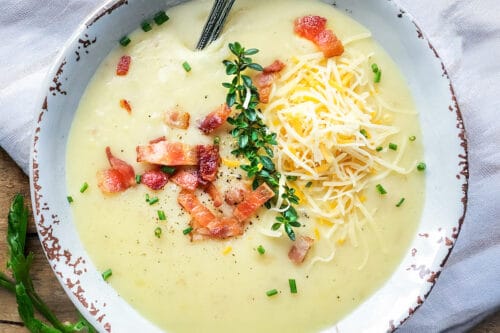 A bowl of creamy Slow Cooker Potato Soup topped with shredded cheese, crispy bacon, fresh herbs, and chives, with a spoon resting inside.
