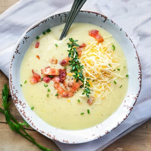 A bowl of creamy Slow Cooker Potato Soup topped with shredded cheese, crispy bacon, fresh herbs, and chives, with a spoon resting inside.