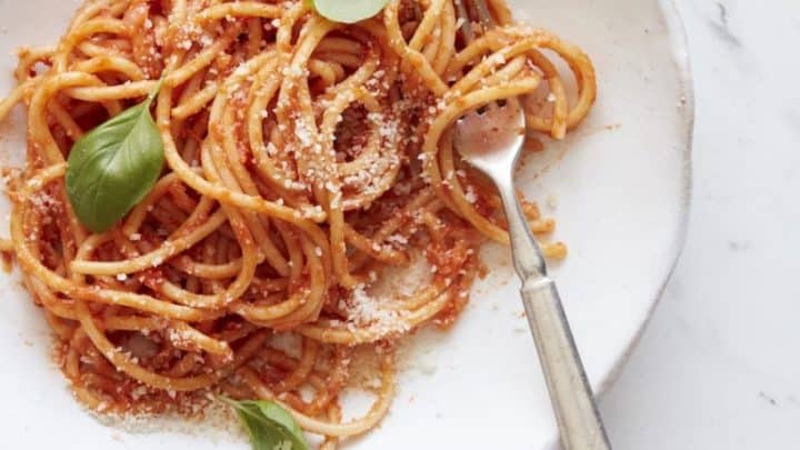 A plate of spaghetti with tomato sauce, grated cheese, and fresh basil leaves—an example of pantry staple recipes—served with a fork on a white marble surface.