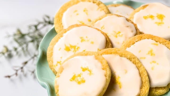 A green plate holds several Lemon Tea Cookies, each topped with white icing and sprinkled with lemon zest; a cup of tea and fresh lemons are in the background.