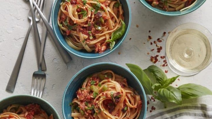 Four bowls of pasta with tomato sauce and herbs, a classic among pantry staple recipes, are arranged on a table with two glasses of white wine, forks, a striped napkin, and fresh basil leaves.