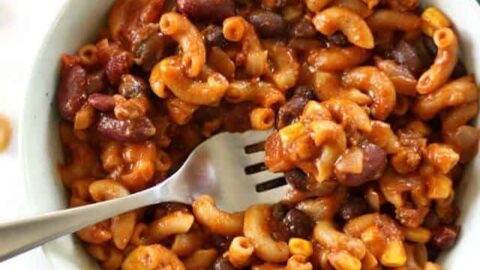 A bowl of macaroni with beans, ground meat, corn, and tomato sauce—a perfect example of pantry staple recipes—sits on a teal cloth napkin, with a fork resting inside.