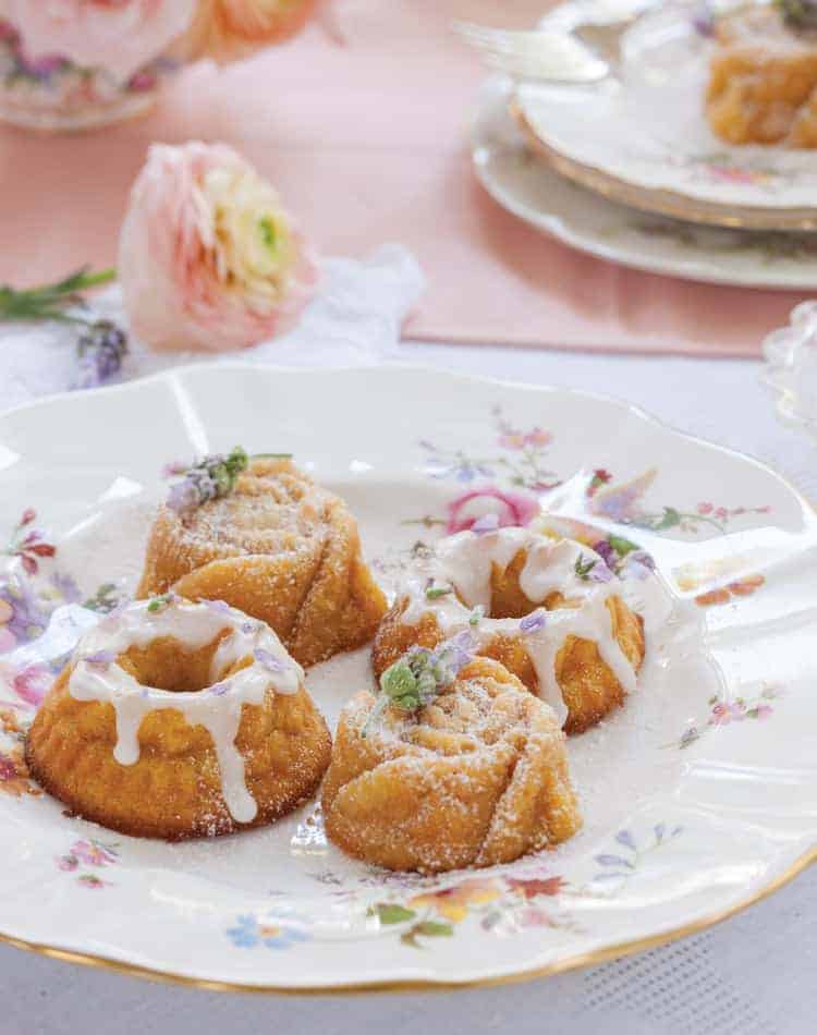 Three small bundt cakes with white icing and powdered sugar are served on a floral-patterned plate, perfect for a spring afternoon tea, with pink flowers and another plate in the background.