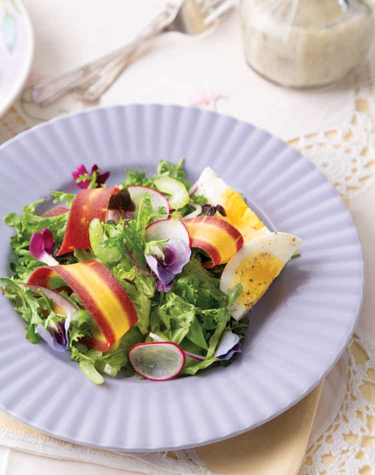 A salad with mixed greens, radish slices, ribboned carrots, hard-boiled egg wedges, and edible flowers on a lavender plate-perfect for a spring afternoon tea.