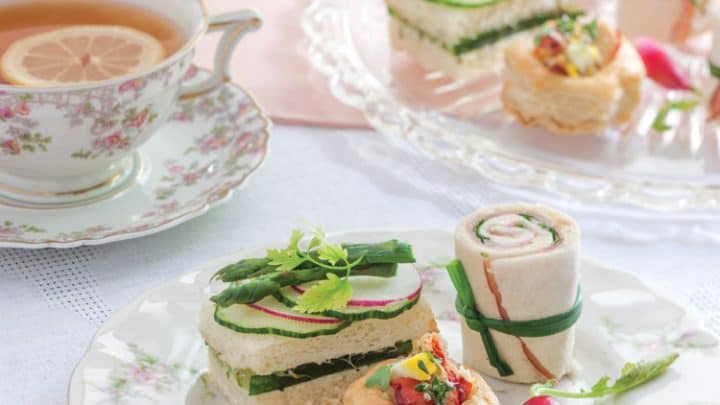 A floral china plate with assorted tea sandwiches, a pastry, and a radish sits next to a teacup with lemon and a tray of more sandwiches on a pink and white table setting, perfect for a spring afternoon tea.