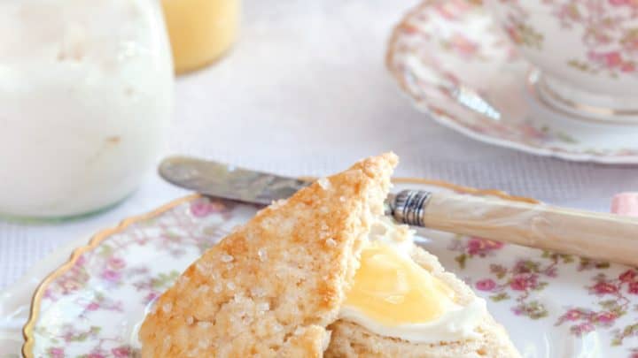 A scone with cream and lemon curd on a floral plate, beside a butter knife, a cup of tea with lemon, and a jar of cream rests on a lace tablecloth—perfect for a spring afternoon tea.
