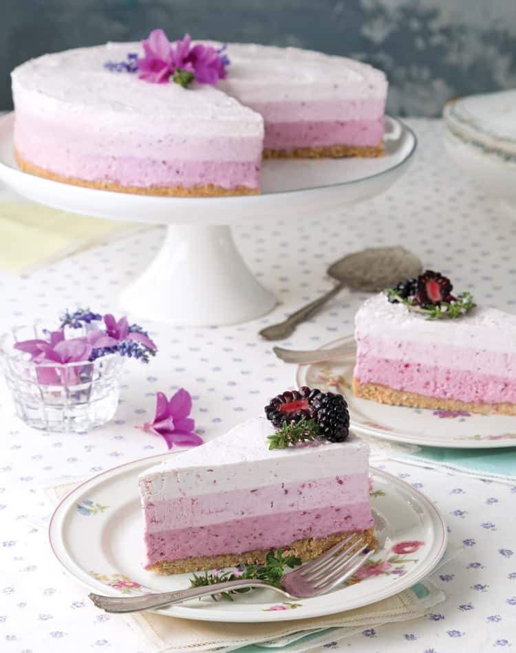 A layered berry mousse cake with a slice served on a plate, garnished with blackberries and edible flowers, sits on a floral tablecloth-perfect for a spring afternoon tea.