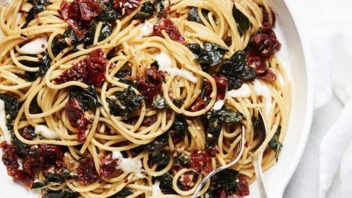 A white bowl filled with spaghetti tossed with sun-dried tomatoes and wilted greens—classic pantry staple recipes—served with a fork and spoon on a white surface.