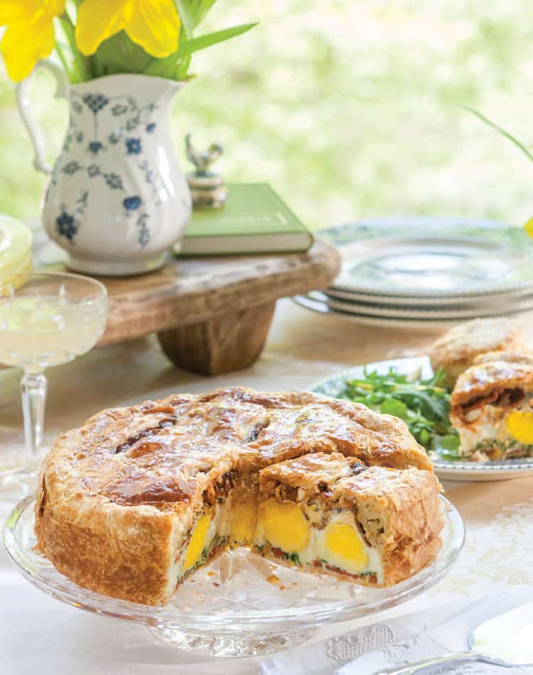 A sliced savory pie with whole eggs inside sits on a glass cake stand, surrounded by plates, glasses, and a vase of yellow flowers-perfect for a spring afternoon tea gathering.