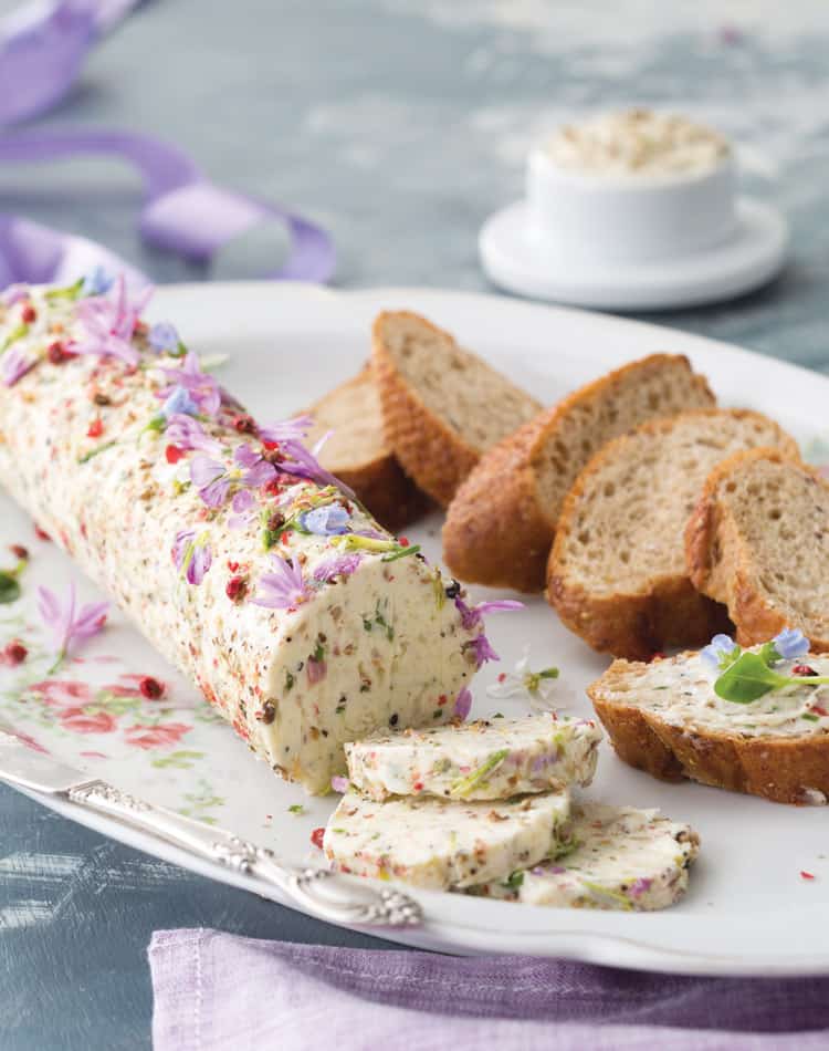 A log of herb and flower-infused butter, partially sliced, is served on a floral plate with several slices of brown bread-perfect for a spring afternoon tea.