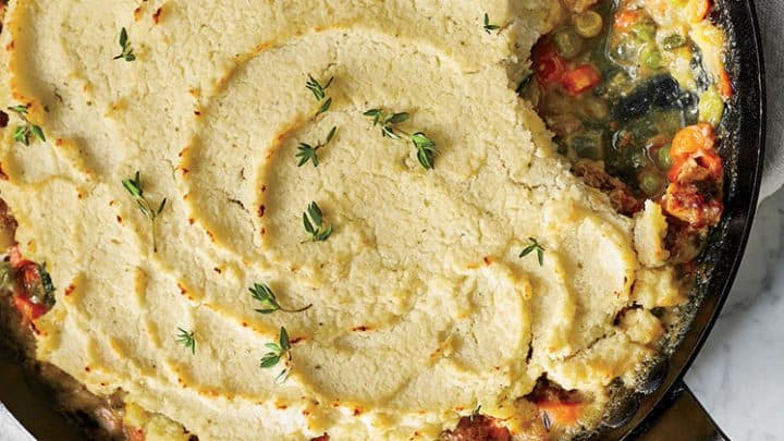 A cast iron skillet filled with shepherd’s pie—one of the classic Healthy Irish Dinners—topped with mashed potatoes and fresh thyme sits on a marble surface next to a glass of white wine.