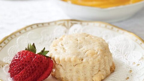 A scone and a strawberry rest on a decorative plate, with a bowl of yellow spread—possibly lemon curd—in the background, perfect for a spring afternoon tea.