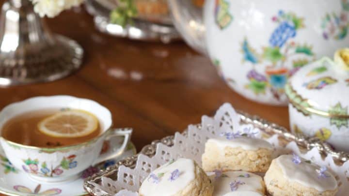 A tea set with floral-patterned cups, saucers, and a teapot evokes a spring afternoon tea, with two cups of tea with lemon slices and a tray of iced scones decorated with purple flowers on a wooden table.