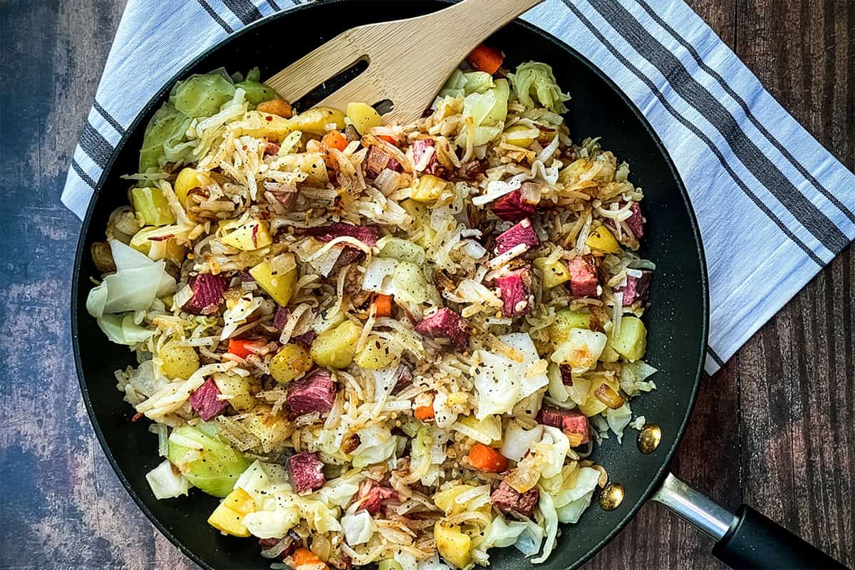 A skillet filled with classic Corned Beef Hash-saut&eacute;ed cabbage, diced potatoes, carrots, and tender corned beef-rests atop a striped kitchen towel.