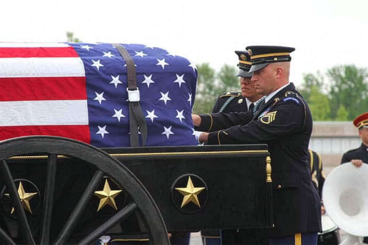 Two uniformed soldiers stand beside a flag-draped casket on a caisson, honoring the fallen during a military funeral ceremony-a solemn reminder of those we remember on Memorial Day.