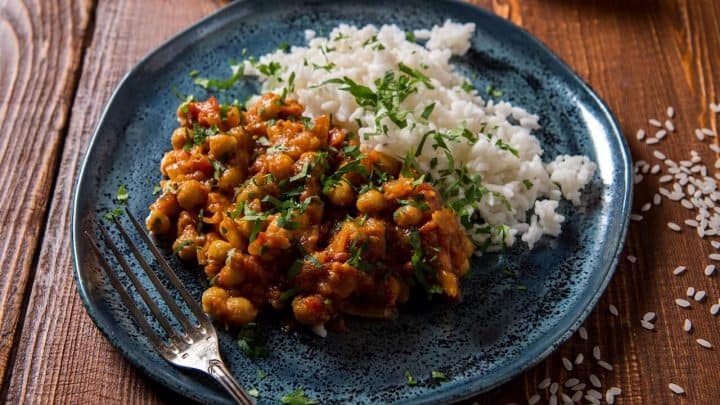 A blue plate with white rice and hearty chickpea curry—classic pantry staple recipes—garnished with chopped herbs, sits on a wooden table with a fork and scattered grains of rice nearby.