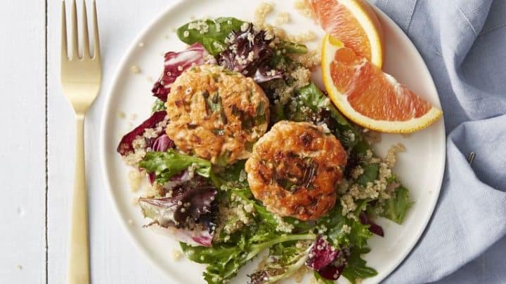 A white plate with two salmon patties on mixed greens and quinoa, garnished with orange slices; a gold fork and blue napkin sit beside the plate—perfect for elegant Mother's Day recipes.