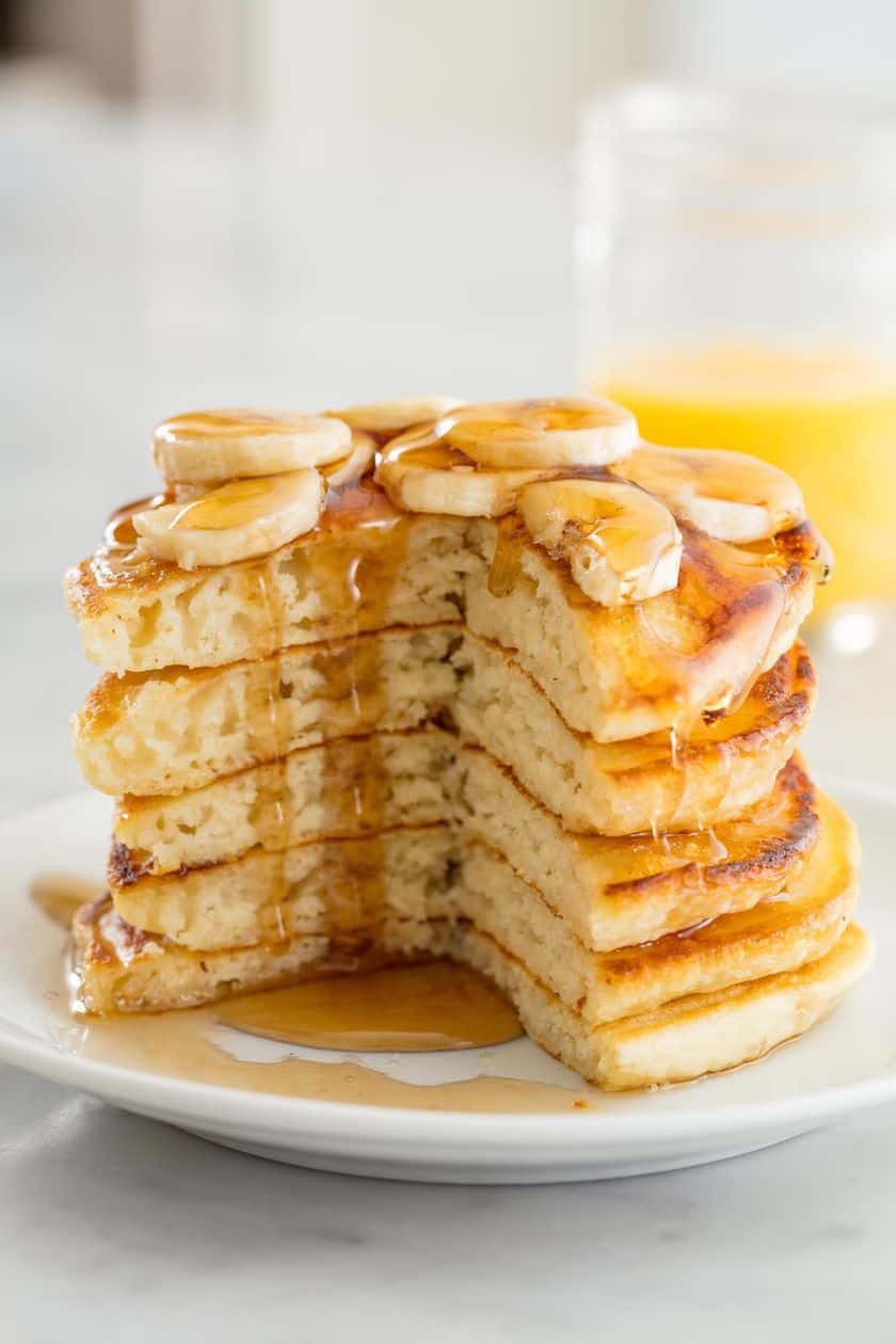 A stack of five pancakes topped with banana slices and syrup, with a large wedge cut out, on a white plate-perfect for adding to your list of Easter Breakfast recipes.