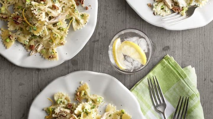 Three plates of bow tie pasta with shredded chicken and herbs sit on a wooden table next to a glass of water with lemon—perfect for sharing as part of your favorite Mother's Day recipes. Two forks and a green-striped napkin add a cozy touch.