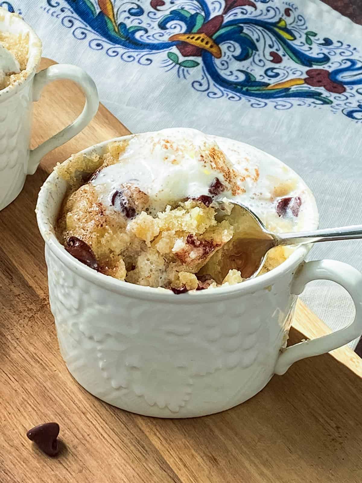 A white mug filled with a warm chocolate chip banana cake topped with melting vanilla ice cream sits on a wooden surface, with a spoon inside and a colorful cloth in the background.