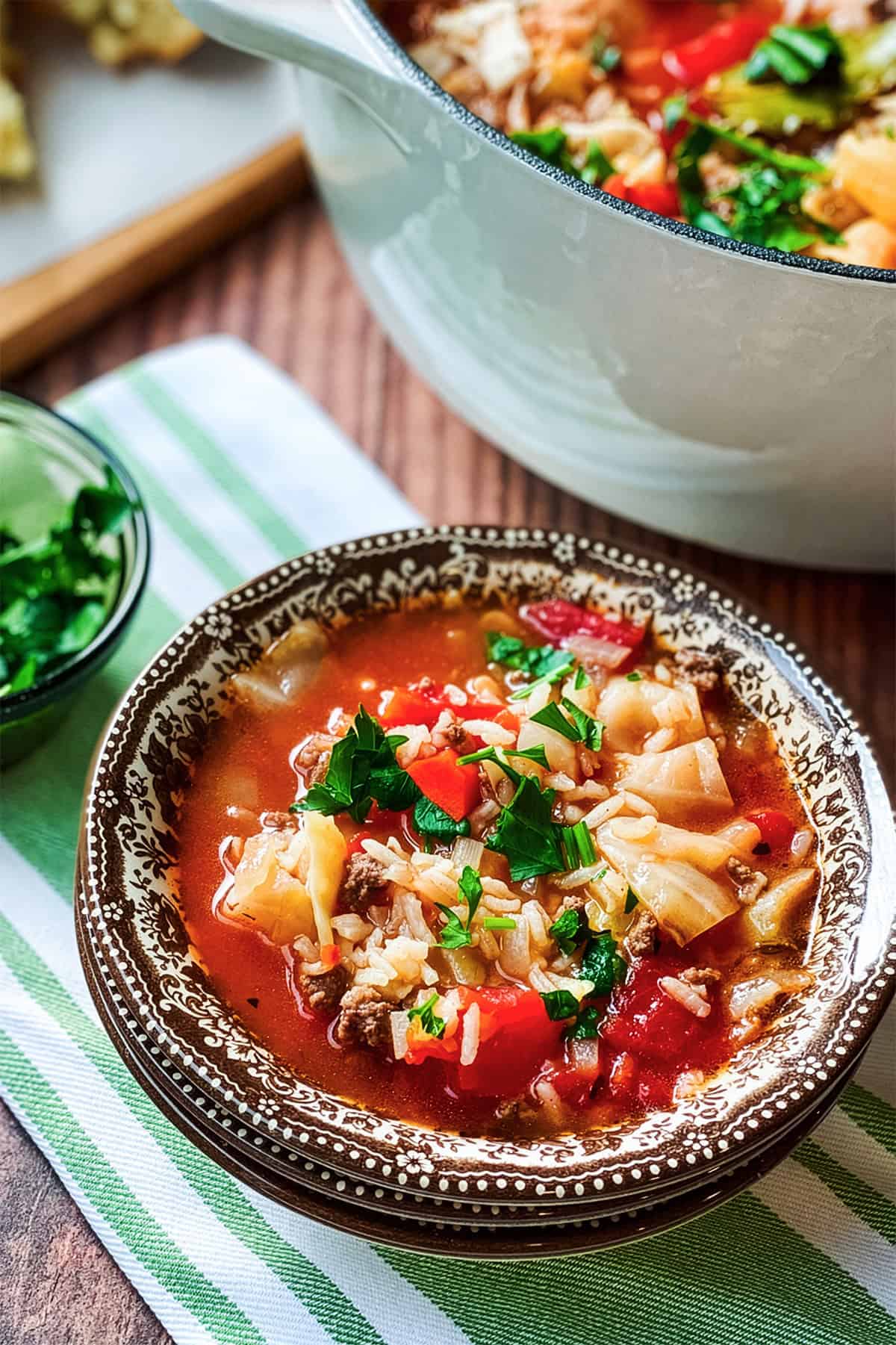 A bowl of cabbage roll soup with ground meat, tomatoes, rice, and parsley, served on a striped cloth with a pot of soup and chopped herbs in the background.