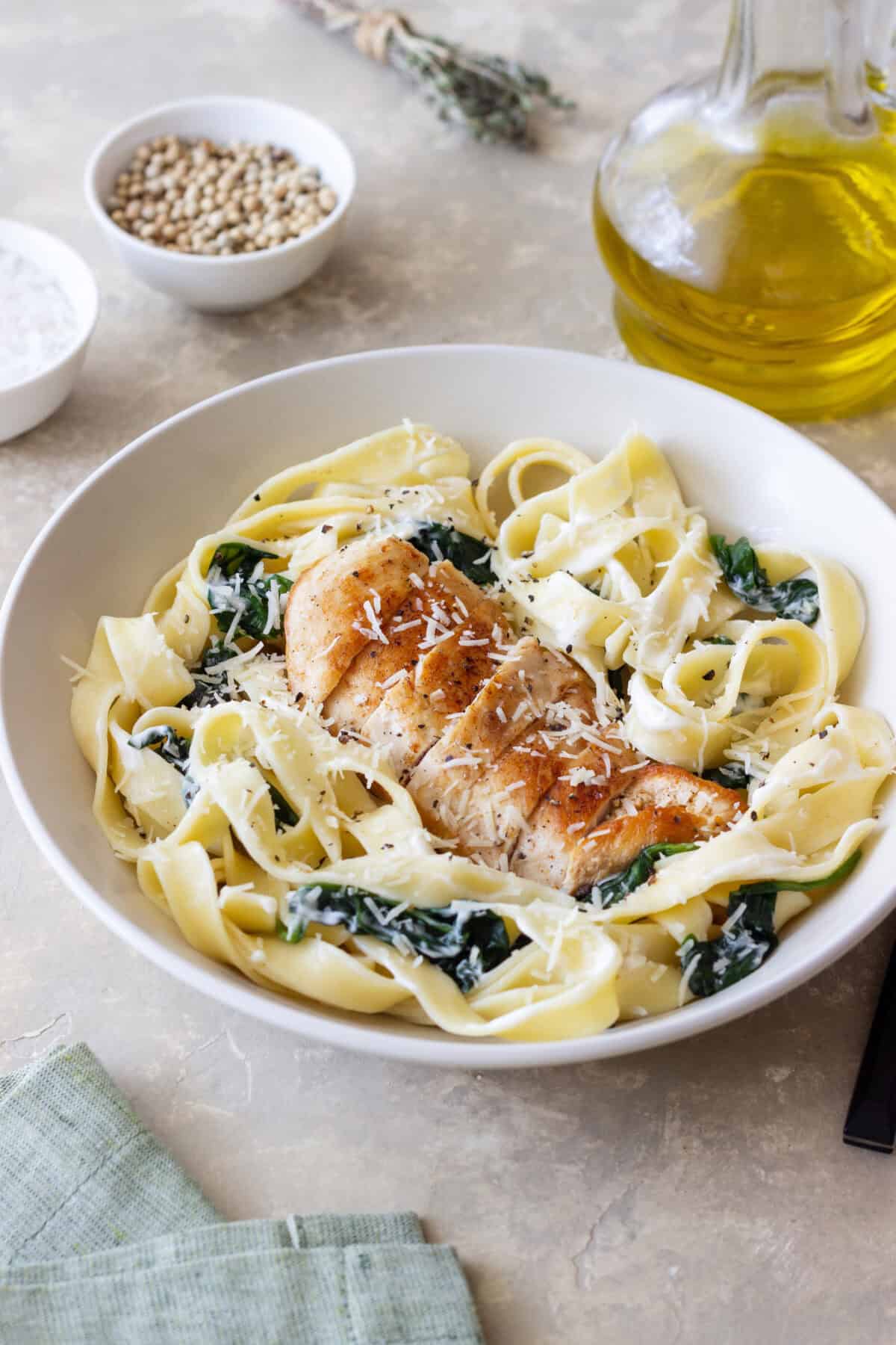 A bowl of chicken fettuccine alfredo with saut&eacute;ed spinach, topped with grated cheese. Olive oil, spices, and a green napkin are arranged around the bowl on a light surface.