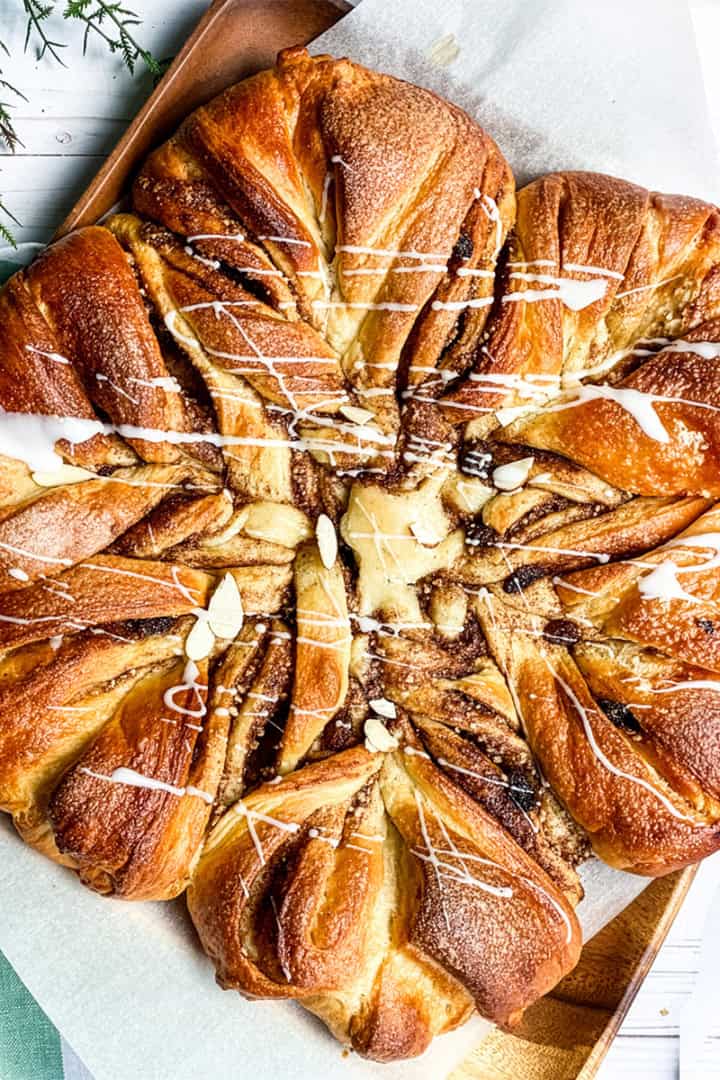Golden-brown star-shaped babka bread with twisted segments, drizzled with white icing, on parchment paper atop a wooden board.