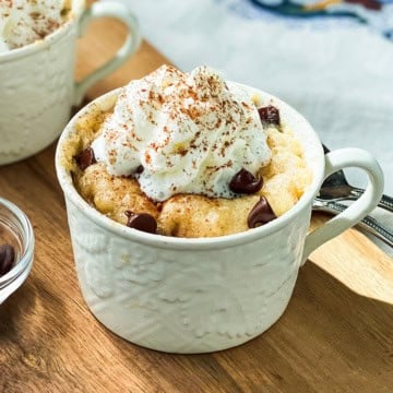 A white mug filled with a chocolate chip banana cake, topped with whipped cream and a sprinkle of cocoa powder, sits on a wooden surface.