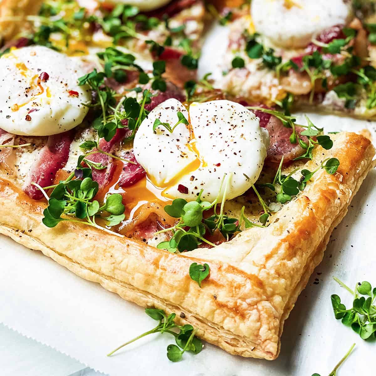 A puff pastry tart topped with poached eggs, bacon, microgreens, and black pepper-an elegant choice among Easter breakfast recipes, served on parchment paper.