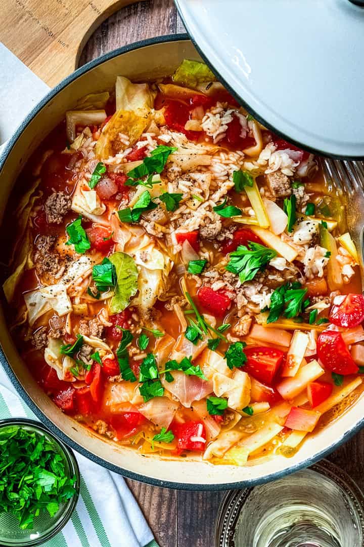 A pot of unstuffed cabbage roll soup with ground meat, tomatoes, rice, and fresh parsley sits on a wooden table beside a glass of white wine, chopped parsley, and a piece of bread.