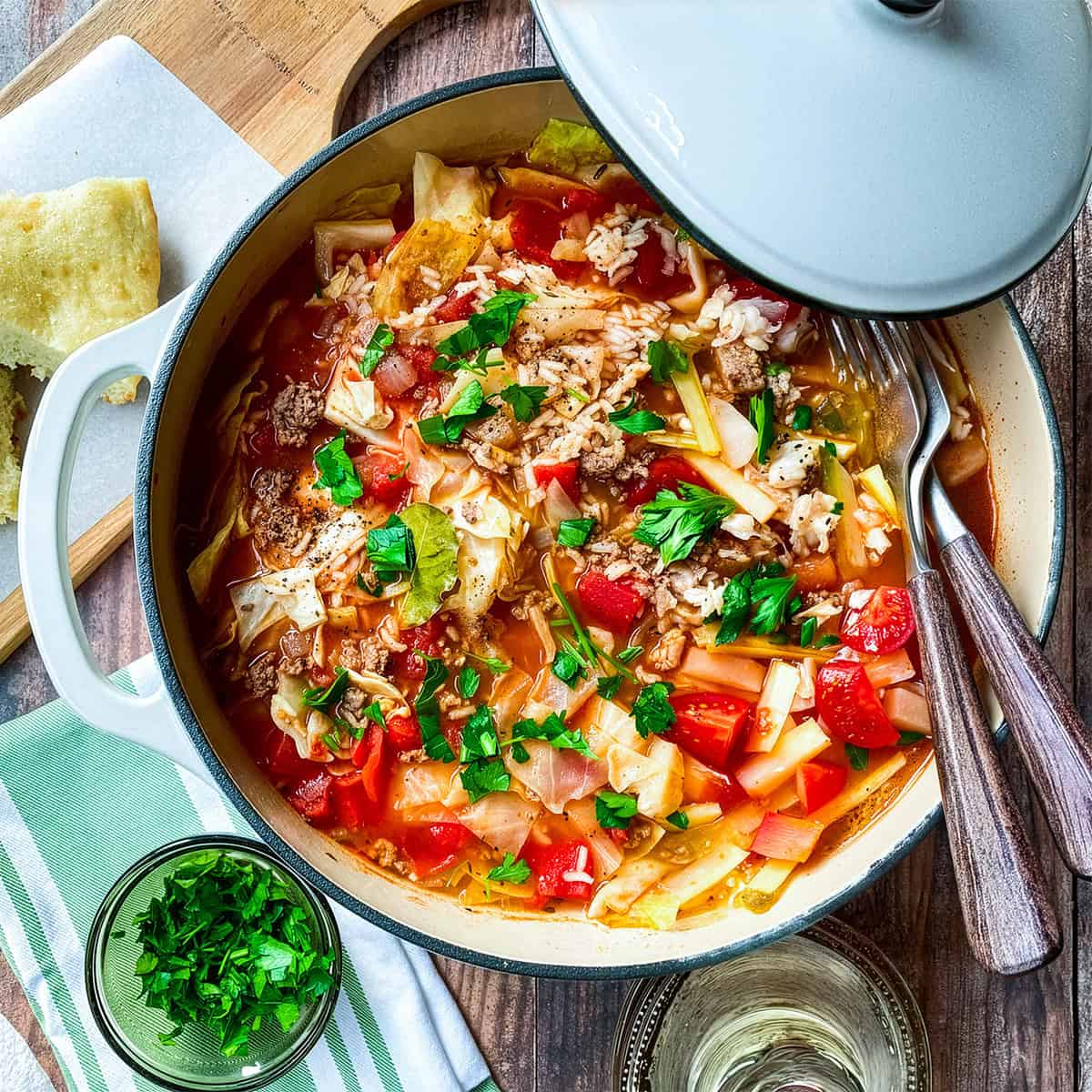 A pot of unstuffed cabbage roll soup with ground meat, tomatoes, rice, and fresh parsley sits on a wooden table beside a glass of white wine, chopped parsley, and a piece of bread.
